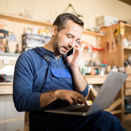 Portrait of mature worker using laptop and speaking by smartphone  in workshop interior, copy space